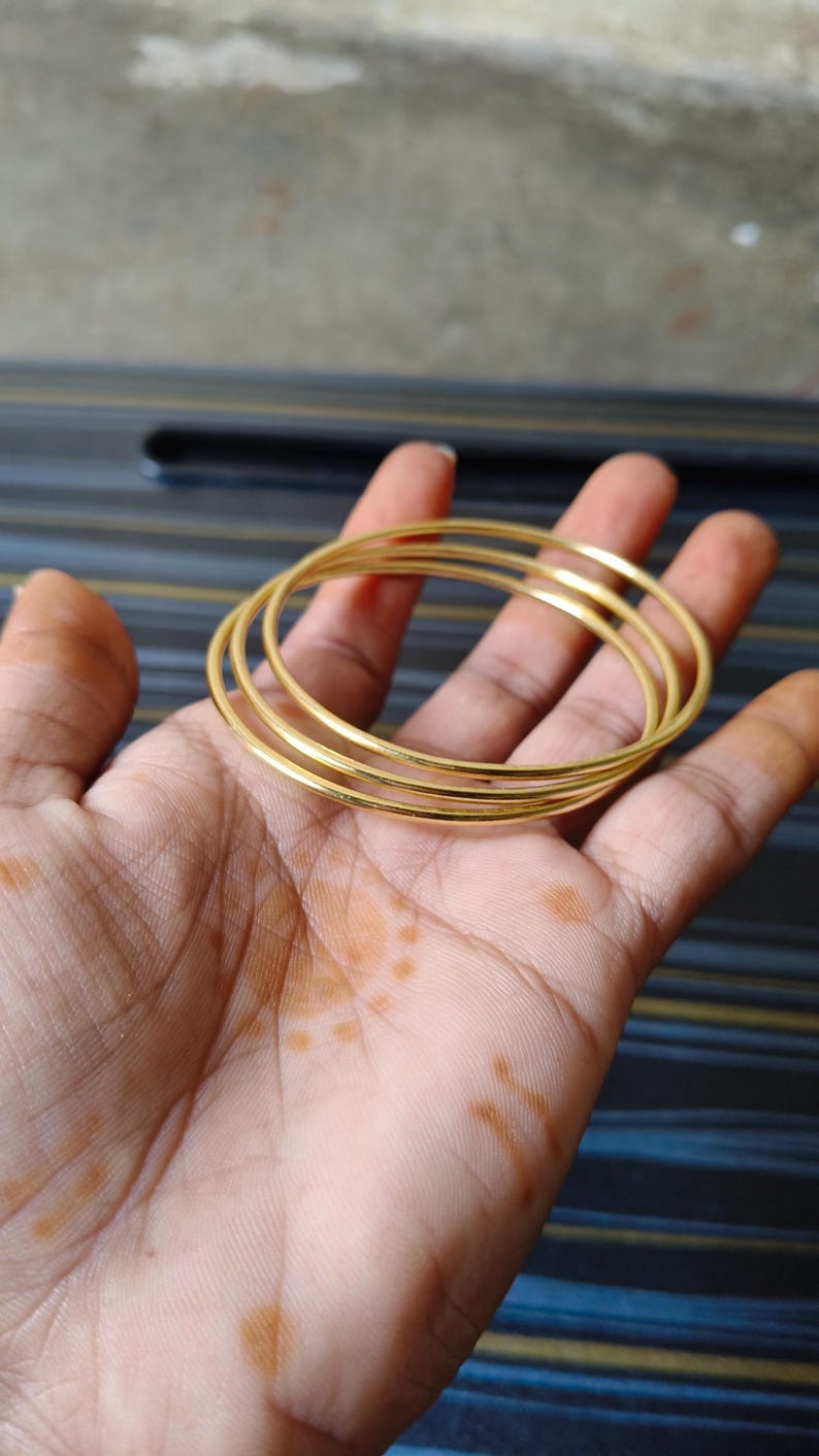 May include: A set of gold-colored bangles, stacked on top of each other, are held in a person's hand. The bangles are thin and have a smooth, reflective surface. The background is a blurred mix of colors.