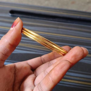May include: A set of gold-colored bangles, held in a hand. The bangles are thin and appear to be made of metal. The background is a dark surface with a striped pattern.