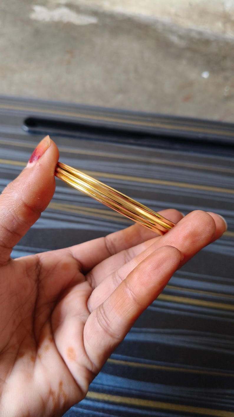 May include: A set of gold-colored bangles, held in a hand. The bangles are thin and appear to be made of metal. The background is a dark surface with a striped pattern.