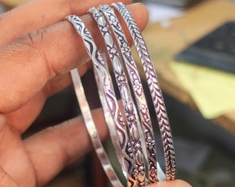 May include: Four silver bangles with intricate floral and geometric patterns. The bangles are held in a hand, showcasing their detailed designs and polished finish. The bangles are a traditional jewelry item.