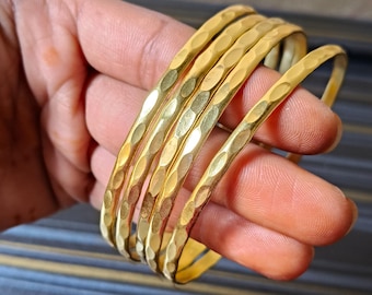 May include: A set of six gold-colored bangles with a hammered texture. The bracelets are held in a hand, showcasing their circular shape and the light reflecting off their surface. The bangles are a warm, metallic color.
