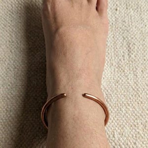 Puede incluir: Una pulsera de tobillo abierta de color cobre rodea un tobillo. La pulsera está hecha de un metal liso y redondeado. El pie y el tobillo descansan sobre una superficie beige texturizada.