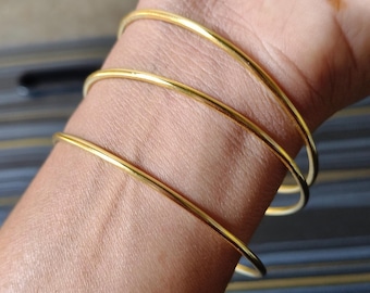 May include: Three gold-colored bangles are stacked on a wrist. The bangles are thin and round, with a smooth, shiny finish. The skin tone is light brown, and the background is blurred.