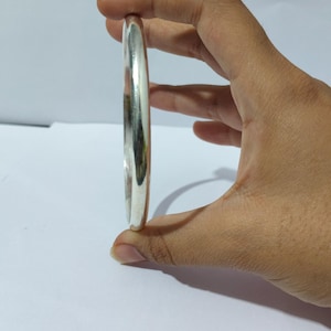 May include: A polished silver bangle bracelet, held in a hand. The bracelet has a smooth, rounded design and reflects light. This piece of jewelry is a simple, elegant accessory.