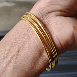 Op de afbeelding: Een set van vier goudkleurige armbanden. De dunne, ronde armbanden zijn op een pols gestapeld en reflecteren licht. De sieraden zijn eenvoudig en elegant, geschikt voor dagelijks gebruik of speciale gelegenheden. Elke armband is ongeveer 2 mm dik.