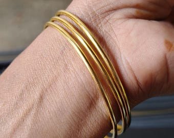 May include: A set of four gold-colored bangles. The thin, round bracelets are stacked on a wrist, reflecting light. The jewelry is simple and elegant, suitable for everyday wear or special occasions.
