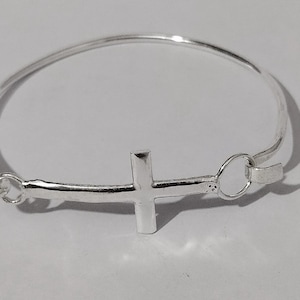 May include: A silver-colored bangle bracelet with a cross charm. The cross is centered on the bracelet and is a solid, three-dimensional design. The bracelet has a simple clasp closure.