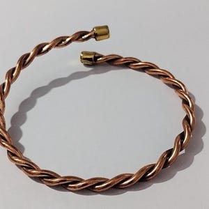 May include: A copper-colored, open cuff bracelet with a twisted rope design. The ends of the bracelet have gold-colored caps. The bracelet is approximately 2.5 inches in diameter.