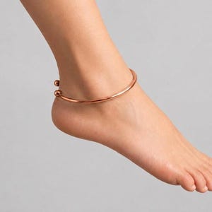 May include: A polished copper-toned metal anklet featuring a simple, open-ended circular band design. The anklet is finished with two small, smooth spherical beads at each terminal end, creating a clean and minimalist look around the ankle.