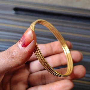 May include: A set of four gold-colored bangles, held in a hand. The bangles are thin and round, with a slightly textured surface. The background is a blurred mix of colors.