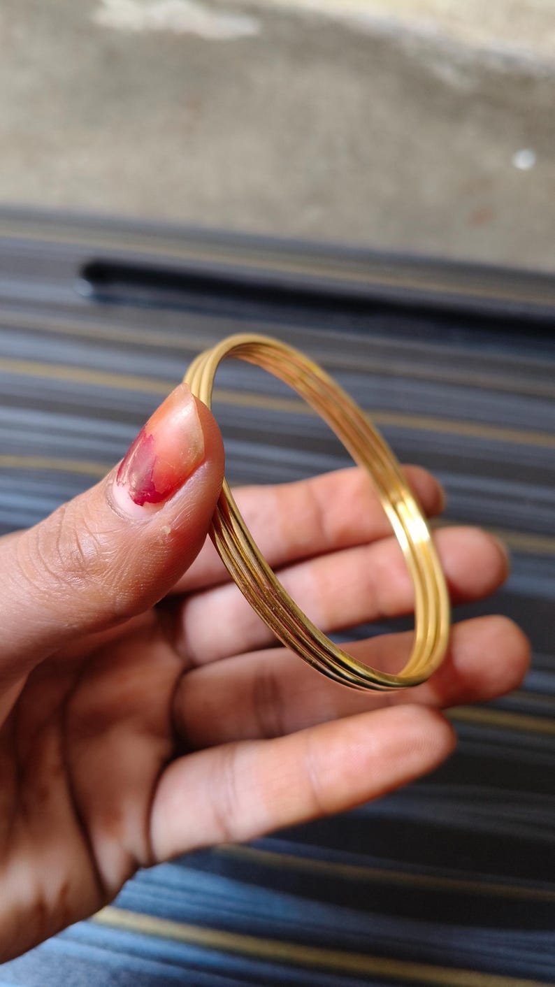 May include: A set of four gold-colored bangles, held in a hand. The bangles are thin and round, with a slightly textured surface. The background is a blurred mix of colors.