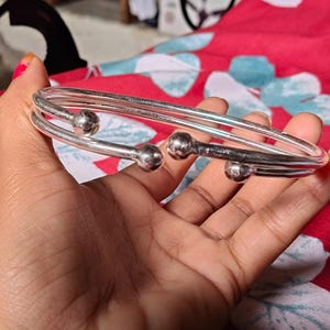 Puede incluir: Una pulsera de brazalete plateada con tres bandas paralelas y pequeñas cuentas redondas plateadas. La pulsera se sostiene en una mano sobre un fondo rojo con estampado floral.