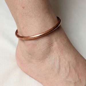 May include: A simple, smooth, circular anklet crafted from polished copper metal. The band features a continuous, solid design with a warm, metallic reddish-brown hue that encircles the ankle, showcasing a clean and unadorned aesthetic.
