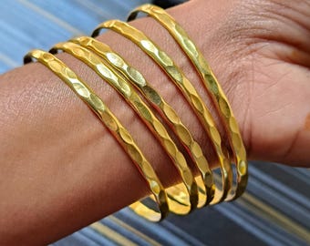 May include: A set of six gold-colored bangles with a hammered texture, worn on a wrist. The bangles are made of a shiny metal and have a slightly irregular surface. The background is a blue and gray striped pattern.