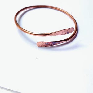 Puede incluir: Un brazalete de color cobre con un diseño abierto. El brazalete tiene una textura martillada en los extremos y una banda lisa y redondeada. Los extremos del brazalete se superponen ligeramente. La joya está sobre un fondo blanco.