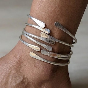 May include: Silver-toned anklet with a layered design. The anklet features multiple curved bands that wrap around the ankle. The ends of the bands have a flattened, teardrop shape. The jewelry has a rustic, handcrafted appearance.