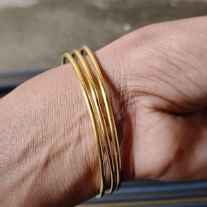 May include: A set of four thin, gold-colored bangles stacked on a wrist. The bangles are smooth and shiny, reflecting light. The background is a mix of neutral tones and a hint of blue stripes.