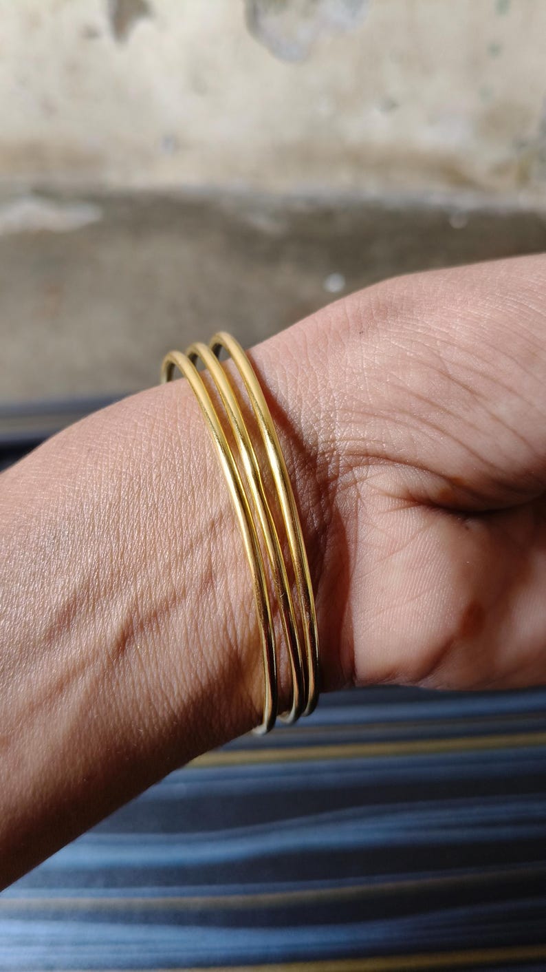 May include: A set of four thin, gold-colored bangles stacked on a wrist. The bangles are smooth and shiny, reflecting light. The background is a mix of neutral tones and a hint of blue stripes.
