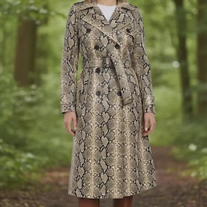 May include: A long trench coat with a snakeskin print in brown, tan, and black. The coat has a double-breasted design and a belt at the waist. The model is wearing brown knee-high boots. The background is a blurred forest.