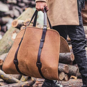 Puede incluir: Un portaleña de cuero marrón con correas y asas de color marrón oscuro. El portaleña está lleno de varios trozos de leña. Una persona sostiene la bolsa.