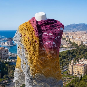 May include: A handmade, open-weave shawl in shades of gray, gold, and deep purple. The shawl is displayed on a mannequin against a backdrop of a city and the sea. The crochet pattern creates a scalloped edge.