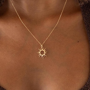 May include: A gold-colored necklace with a sun pendant. The sun pendant has a circular center with rays extending outward. The necklace is worn on a person with brown skin, wearing a brown halter top.