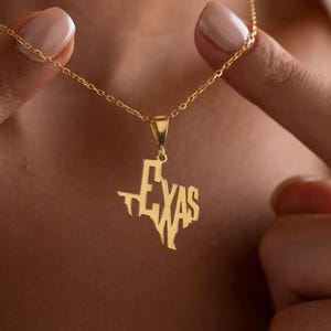 May include: A gold-colored necklace featuring a pendant in the shape of the state of Texas. The word "TEXAS" is cut out within the pendant. The necklace is being held up by two hands.