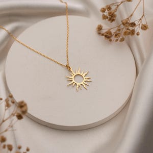 May include: A gold-colored necklace featuring a sun pendant. The sun pendant has a circular center with radiating rays. The necklace is displayed on a white, circular surface with a white, silky background. Dried flowers are in the background.