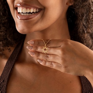 May include: A gold-colored necklace featuring a sun pendant. The sun pendant has a detailed design with rays extending outwards. The necklace is worn around the neck and is held in the hand.