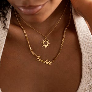 May include: Two gold necklaces. The top necklace has a sun pendant. The bottom necklace has the name "Scarlett" in cursive. The necklaces are layered and worn on a person.