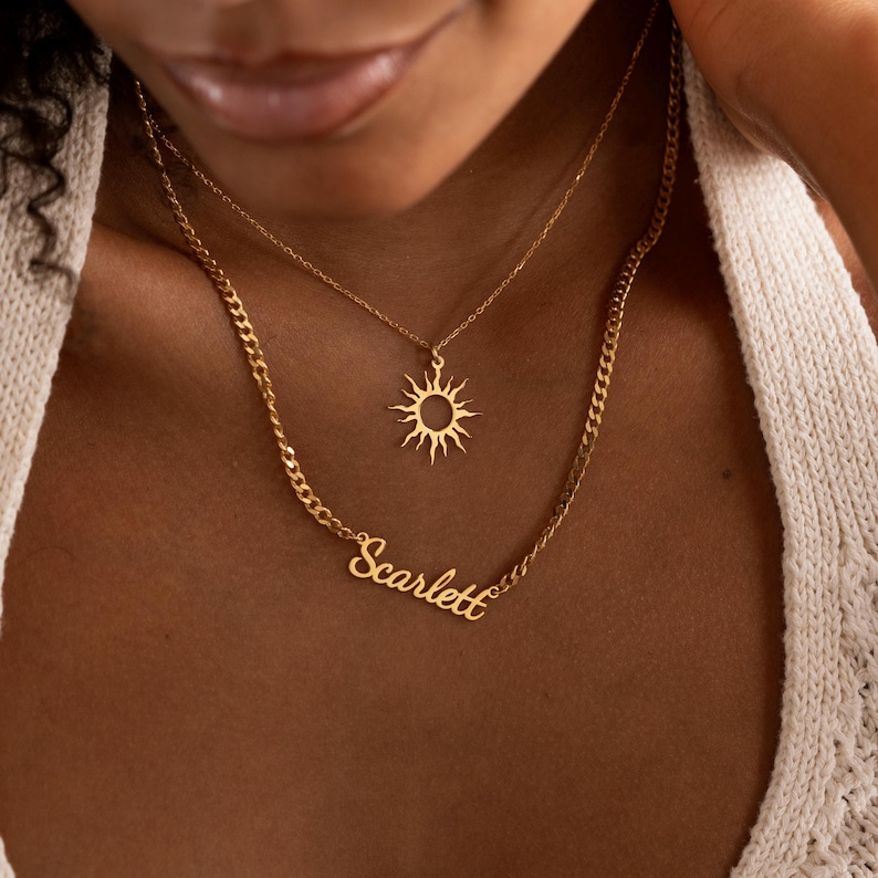May include: Two gold necklaces. The top necklace has a sun pendant. The bottom necklace has the name "Scarlett" in cursive. The necklaces are layered and worn on a person.