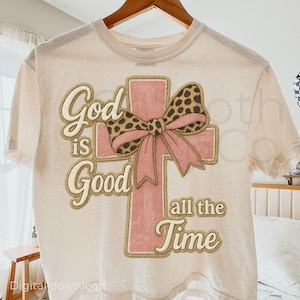 Puede incluir: Camiseta color crema con un diseño de cruz rosa. La cruz está adornada con un lazo con estampado de leopardo. Las palabras "God is Good all the Time" están impresas en fuente dorada.