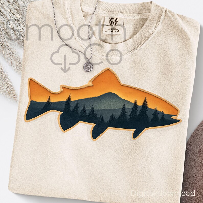 May include: Cream-colored Comfort Colors t-shirt featuring a fish-shaped design with a mountain and forest scene in shades of orange, green, and blue. A silver necklace with a circular pendant is draped over the shirt. The shirt has a tag that says "Comfort Colors".