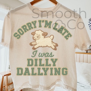 Puede incluir: Camiseta color crema con el texto "SORRY I'M LATE" sobre una oveja de dibujos animados y el texto "I WAS DILLY DALLYING" en verde. La camiseta tiene mangas cortas y cuello redondo.