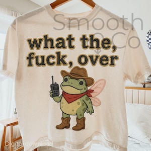 May include: A cream-coloured t-shirt featuring the phrase "what the fuck, over" in a textured brown font. The design includes a cartoon frog in a cowboy hat, red bandana, and boots, holding a walkie-talkie, with fairy wings.