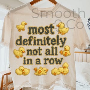 May include: Cream-colored t-shirt with the phrase "most definitely not all in a row" in dark gray letters. Yellow cartoon duck graphics are scattered around the text. The shirt is hanging on a wooden hanger.