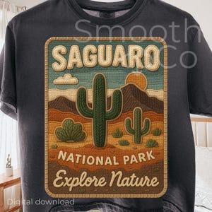 May include: Dark gray t-shirt featuring a rectangular graphic with a desert landscape. The graphic includes the text "SAGUARO NATIONAL PARK" and "Explore Nature" with images of cacti, mountains, and a setting sun.