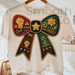 May include: A cream-coloured t-shirt with a large bow design. The bow is made up of red, black, and green sections with gold details, including a fist, star, and floral patterns. The text "Smooth Co" is visible on the shirt.