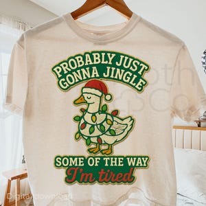 May include: Cream-colored t-shirt with a Christmas-themed graphic. The graphic features a white duck wearing a Santa hat and Christmas lights, with the text "Probably Just Gonna Jingle Some of the Way I'm Tired."