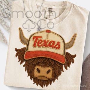 May include: Cream-colored t-shirt featuring a brown and tan Highland cow wearing a baseball cap with "Texas" in red lettering. The shirt has a Comfort Colors tag and a silver necklace.