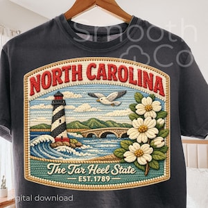 May include: Dark gray t-shirt featuring a detailed embroidered design of North Carolina. The design includes a lighthouse, bridge, seagull, and dogwood flowers, with the text "North Carolina" and "The Tar Heel State".