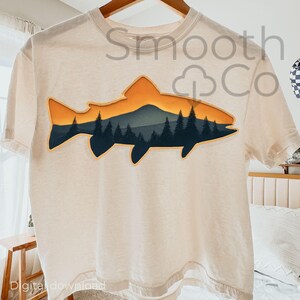 May include: Cream-colored t-shirt featuring a graphic of a fish silhouette with a mountain and forest scene inside. The design includes shades of orange, blue, and green. The text "Smooth Co" is visible on the shirt.