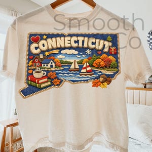May include: Cream-colored t-shirt featuring a detailed graphic of the state of Connecticut. The design includes the word "Connecticut" in a blue, stitched-style border, with images of sailboats, autumn foliage, and a coffee cup.