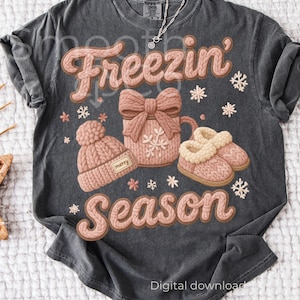 May include: Dark gray t-shirt with a winter-themed graphic. The design features the words "Freezin' Season" in a textured, light brown font, along with illustrations of a knit hat, mug, and slippers. Snowflakes and the words "Digital download" are also visible.