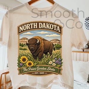May include: A cream-coloured t-shirt with a detailed graphic of a bison in a field. The design includes the words "North Dakota" and "The Peace Garden State", along with sunflowers, daisies, and the text "EST. 1889".