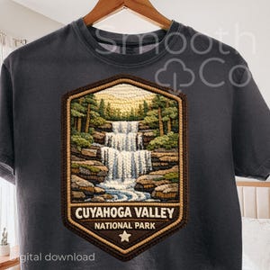 Puede incluir: Camiseta gris oscuro con un parche detallado que representa una escena de cascada del Parque Nacional Cuyahoga Valley. El parche incluye árboles, rocas y el nombre del parque. La camiseta está colgada en una percha de madera.