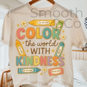 May include: Cream-colored t-shirt with a colorful graphic design. The design features the words "COLOR the world WITH KINDNESS" in large, multi-colored letters, surrounded by crayon illustrations and floral accents. The shirt also includes the text "you are loved", "you matter", "believe in yourself", "you are enough", and "you are smart".