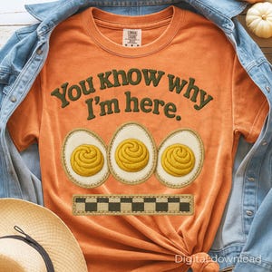 Puede incluir: Camiseta naranja con el texto "You know why I'm here." y tres diseños de huevos bordados. Debajo de los huevos hay un patrón de cuadros. La camiseta está doblada y sobre una chaqueta vaquera y un sombrero de paja.