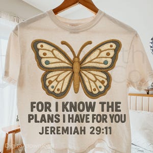 May include: Cream-colored t-shirt featuring a large butterfly design in shades of brown, tan, and teal. The text "FOR I KNOW THE PLANS I HAVE FOR YOU JEREMIAH 29:11" is printed below the butterfly. The shirt is hanging on a wooden hanger.