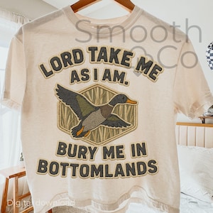 以下が含まれることがあります： 「LORD TAKE ME AS I AM. BURY ME IN BOTTOMLANDS」のテキストが入ったクリーム色のTシャツ。テキストの下には、六角形の中にアヒルのグラフィックが配置されています。アヒルは飛行中で、緑、茶色、オレンジ色のディテールがあります。